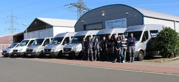 Eine Gruppe von Menschen steht vor einer Reihe von weißen Lieferwagen vor einem Industriegebäude.