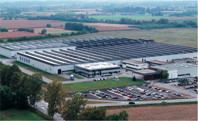 Ein großes Industrie- oder Logistikgelände mit mehreren langgestreckten Gebäuden, umgeben von Parkplätzen und ländlicher Landschaft.