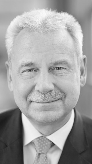 Ein Mann mit grauem Haar, Anzug und Krawatte schaut ernst in die Kamera.