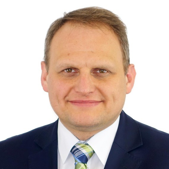 Ein Mann mit kurzen blonden Haaren, lächelnd, trägt einen dunkelblauen Anzug mit einer gestreiften Krawatte.