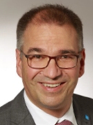 Ein Mann mit Brille und einem Lächeln auf dem Gesicht, der einen Anzug trägt und ein blaues Abzeichen am Revers hat.