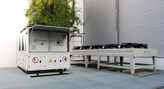 Ein Transportwagen mit einem Container steht neben einem leeren Metallgestell vor einem Gebäude mit einer großen, strukturierten Fassade.