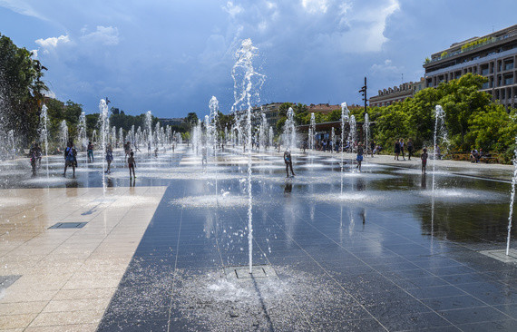 Eine öffentliche Wasserfontänenanlage mit sprudelnden Wasserstrahlen und Menschen, die sich im Hintergrund aufhalten.