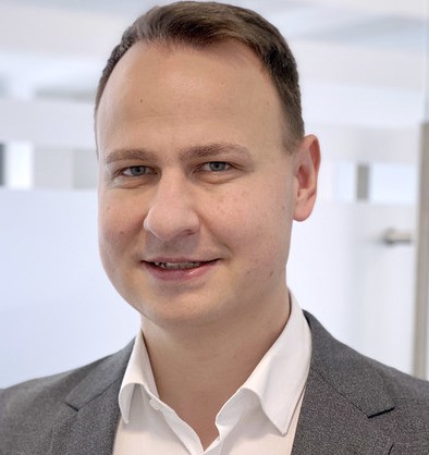 Ein Mann mit dunklem Haar und Bart trägt ein weißes Hemd und einen dunklen Blazer, steht vor einem unscharfen Hintergrund.