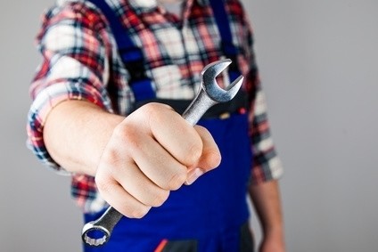 Eine Person in einer karierten Hemdbluse und einer Schürze hält einen Schraubenschlüssel in die Kamera.