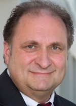 Ein Mann mit dunklem Haar und Bart trägt einen dunklen Anzug und ein weißes Hemd.