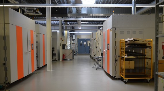 Ein langer, heller Lager- oder Laborgang mit orangefarbenen Schranktüren, Metallregalen und technischen Geräten.