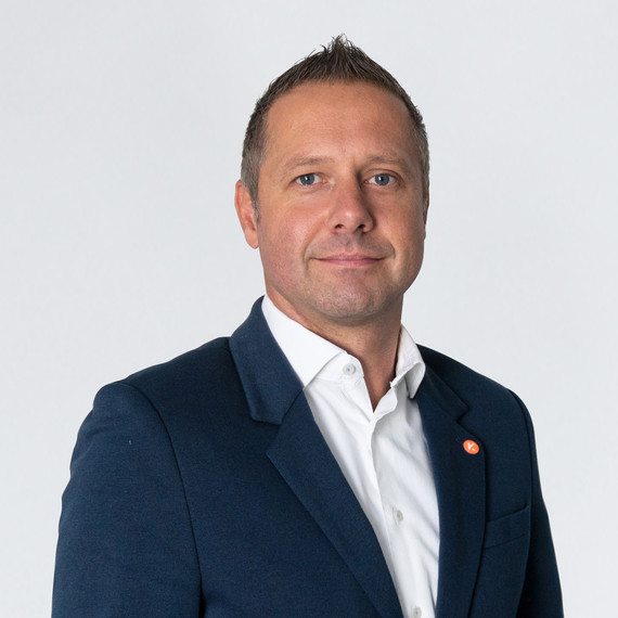 Ein Mann mit kurzem Haar trägt einen dunklen Anzug und ein weißes Hemd, wobei er direkt in die Kamera blickt.