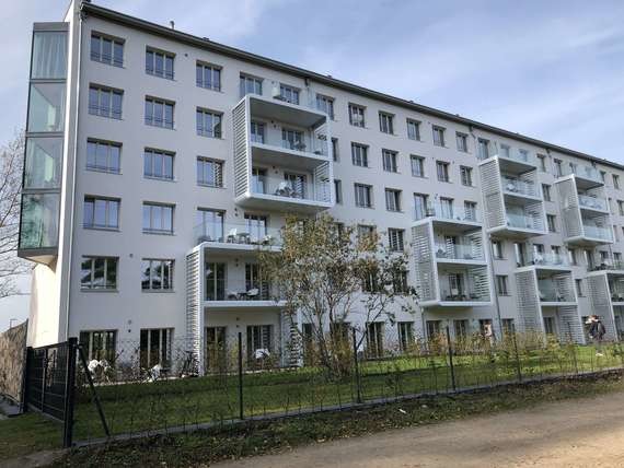 Ein modernes Mehrfamilienhaus mit mehreren Stockwerken und Fensterreihen steht hinter einem Zaun und Grünflächen.