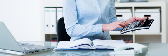 Eine Person sitzt an einem Schreibtisch, tippt auf einer Tastatur und hat vor sich ein geöffnetes Buch sowie einen Laptop.
