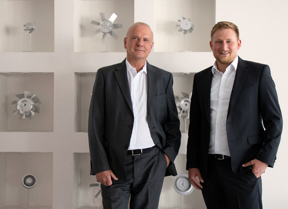 Zwei Männer in Anzügen stehen nebeneinander vor einer weißen Wand mit dekorativen Elementen.