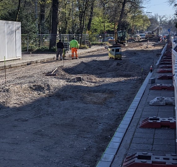 Eine Baustelle mit Arbeiter*innen in Warnwesten, die auf einer Straße mit geparkten Autos und einem teilweise fertiggestellten Gehweg arbeiten.