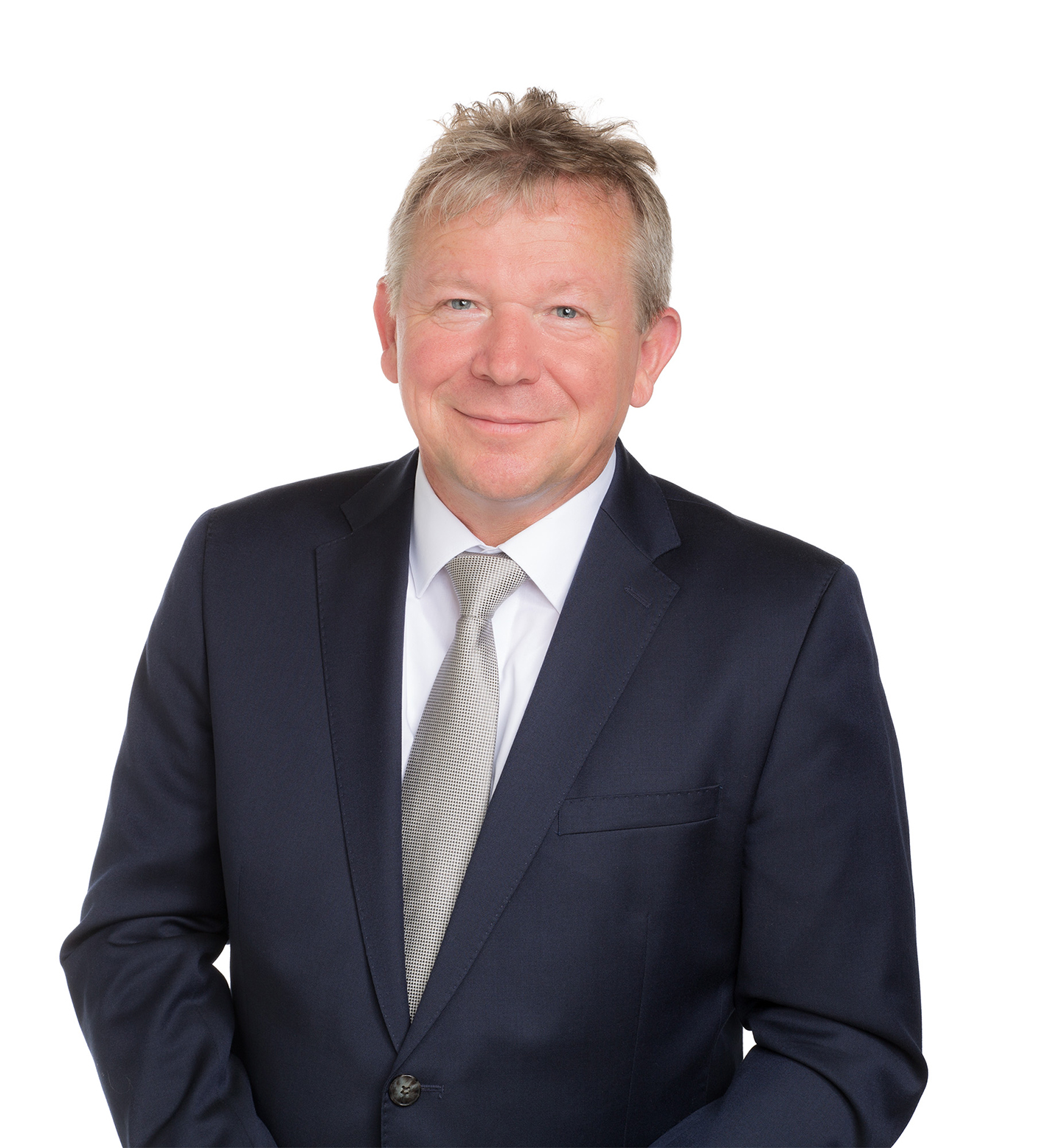 Ein Mann mit kurzen blonden Haaren trägt einen dunkelblauen Anzug und eine graue Krawatte, lächelt und steht vor einem weißen Hintergrund.