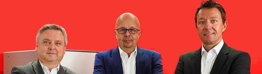 Drei Männer in Anzügen stehen nebeneinander vor einem roten Hintergrund.