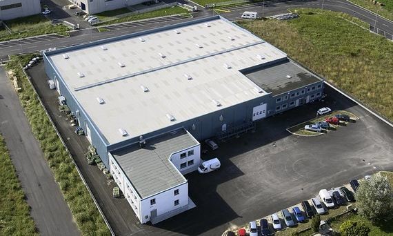 Ein Luftbild eines großen, flachen Industriegebäudes mit einem Parkplatz davor, umgeben von Grünflächen.