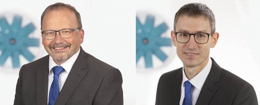 Zwei Männer in Anzügen und Krawatten mit einem Logo im Hintergrund.