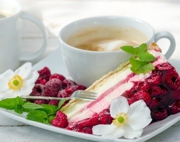 Eine Tasse Kaffee mit Schaum steht neben einem Teller mit einer Scheibe Kuchen, belegt mit Himbeeren und Blüten.