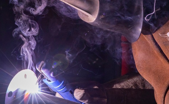 Eine Person im Schweißschutzhelm führt eine Schweißarbeit aus, wobei Funken und Rauch sichtbar sind.