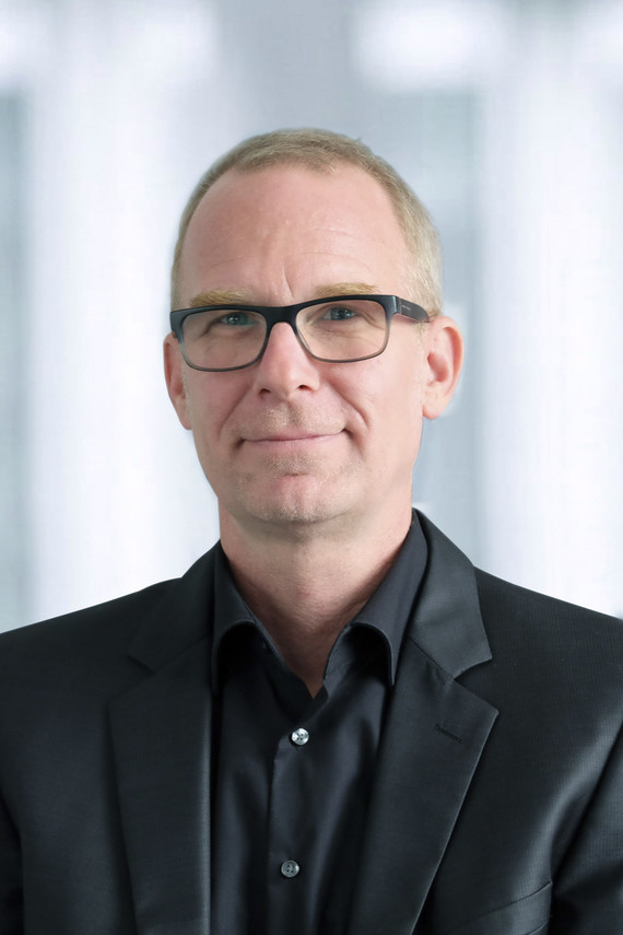 Ein Mann mit kurzen blonden Haaren trägt ein schwarzes Hemd und einen dunklen Anzug, vor einem unscharfen Hintergrund.