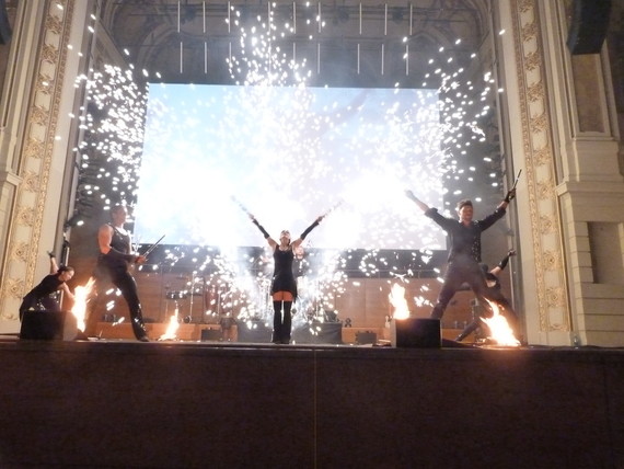 Drei Personen stehen auf einer Bühne mit Feuerwerk im Hintergrund, während sie ihre Arme in die Luft strecken.