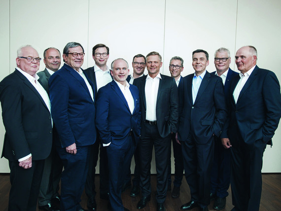 Eine Gruppe von neun Männern in Anzügen steht nebeneinander in einer Reihe.