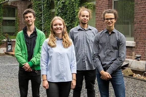 Vier Personen stehen im Freien nebeneinander, lächelnd und in lässiger Kleidung.