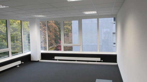 Ein leeres Büro mit großen Fenstern, die auf einen Park mit Bäumen blicken, und einer schwarzen Heizung an der Wand.