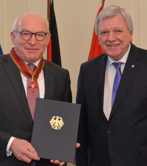 Zwei Männer in Anzügen stehen nebeneinander, einer hält eine Urkunde mit dem deutschen Bundesadler.