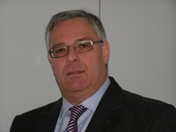 Ein Mann im Anzug und Krawatte sitzt vor einem neutralen Hintergrund.