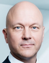 Ein Mann mit kahlen Kopf und blauen Augen schaut ernst in die Kamera.