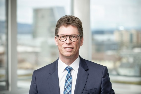 Ein Mann mit Brille, Anzug und Krawatte steht vor einem Fenster mit städtischem Hintergrund.