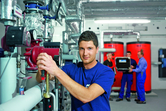 Ein Mann in blauem Hemd arbeitet an einer Rohrleitung in einer technischen Anlage, während im Hintergrund weitere Personen beschäftigt sind.