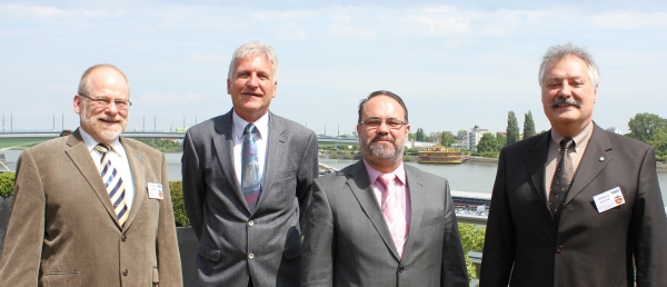 Vier Männer in Anzügen stehen nebeneinander vor einem Hintergrund mit Wasser und einem Boot.