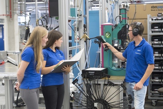 Drei Personen in blauen Hemden stehen in einer industriellen Umgebung und diskutieren, wobei eine Person ein Tablet hält und eine andere ein Messgerät bedient.