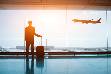 Ein Mann mit Koffer steht im Flughafen und blickt auf ein abfliegendes Flugzeug.