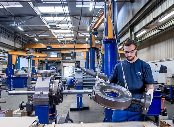 Ein Mann in Arbeitskleidung hält ein großes Metallteil in einer industriellen Werkstatt.