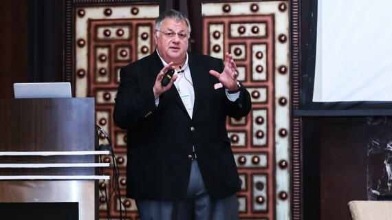 Ein Mann in einem Anzug steht auf einer Bühne und spricht, während er gestikuliert.