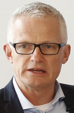 Ein Mann mit grauem Haar und Brille trägt ein dunkles Hemd und ein hellblaues Hemd darunter.