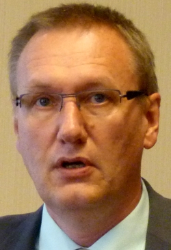 Ein Mann mit Brille und Anzug spricht in einem formellen Setting.