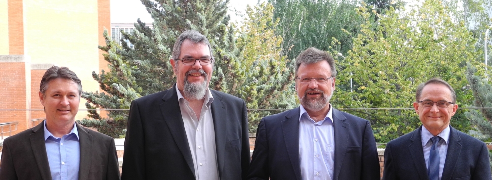 Vier Männer stehen nebeneinander im Freien vor einem Gebäude mit Bäumen im Hintergrund.