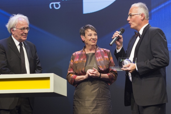 Drei Personen stehen auf einer Bühne, wobei eine Frau in der Mitte einen Preis entgegennimmt, während zwei Männer links und rechts von ihr stehen.