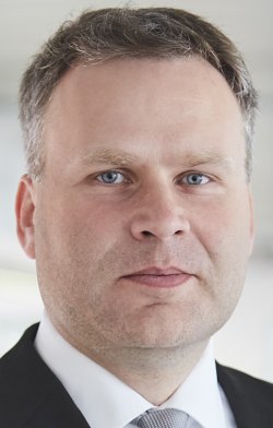 Ein Mann mit kurzen dunklen Haaren trägt einen dunklen Anzug und ein weißes Hemd mit Krawatte.