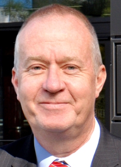 Ein Mann mit grauem Haar und Bart trägt einen dunklen Anzug und ein weißes Hemd.