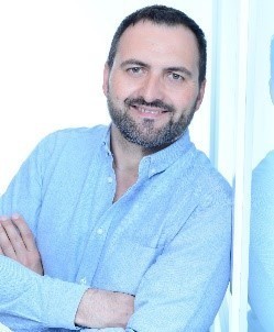 Ein Mann mit dunklem Haar und Bart trägt ein hellblaues Hemd und steht mit verschränkten Armen.