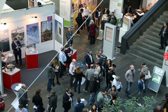 Eine Menschenmenge bewegt sich in einem belebten Ausstellungs- oder Kongressraum mit Ständen, Plakaten und Treppen im Hintergrund.