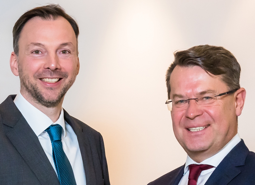 Zwei Männer in Anzügen und Krawatten lächeln in die Kamera.