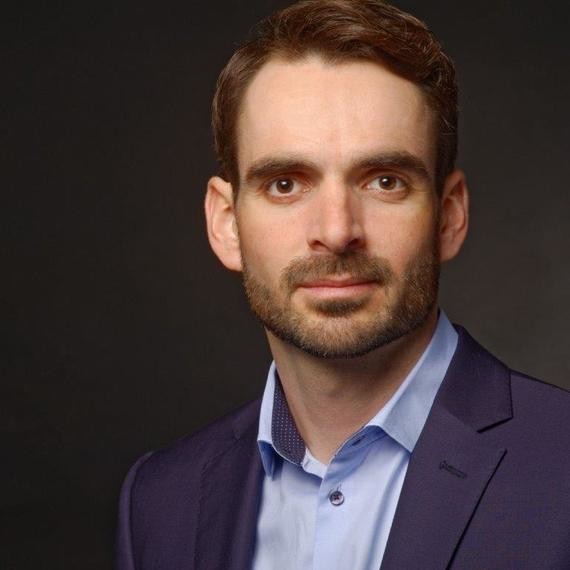 Ein Mann mit dunklem Haar und Bart trägt einen blauen Anzug mit einem hellblauen Hemd und steht vor einem dunklen Hintergrund.