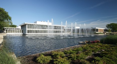 Ein modernes Gebäude mit einer großen Wasserfläche und Fontänen im Vordergrund, umgeben von grüner Vegetation unter blauem Himmel.