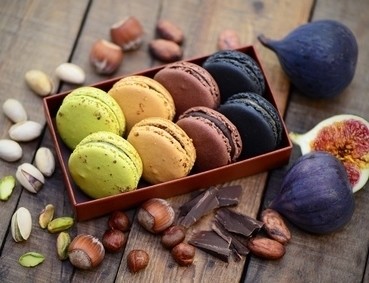 Eine Auswahl an farbenfrohen Macarons in einer Holzschachtel, umgeben von Nüssen, Feigen und Schokoladenstücken auf einem Holztisch.