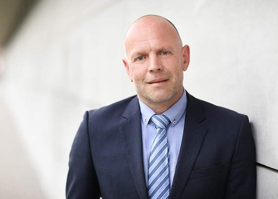 Ein Mann mit kahlköpfigem Haar trägt einen dunklen Anzug mit gestreifter Krawatte und steht vor einem unscharfen Hintergrund.